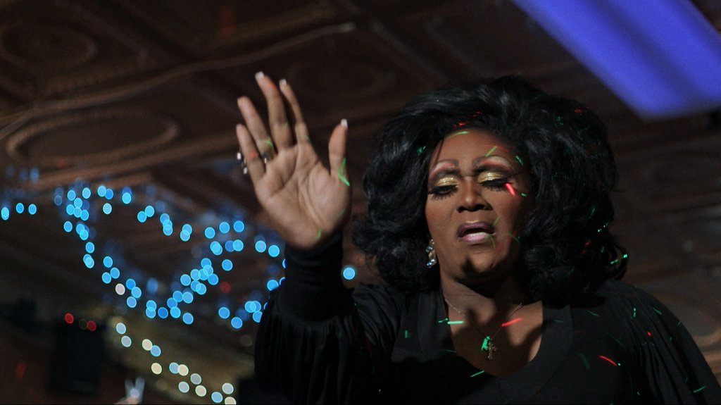 A Black femme person played by Charnay Malletti Cassadine stands with hand outstretched