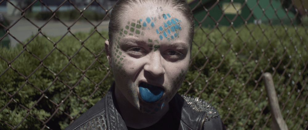 person with blue and green face paint stands in front of a fence with their tongue stuck out