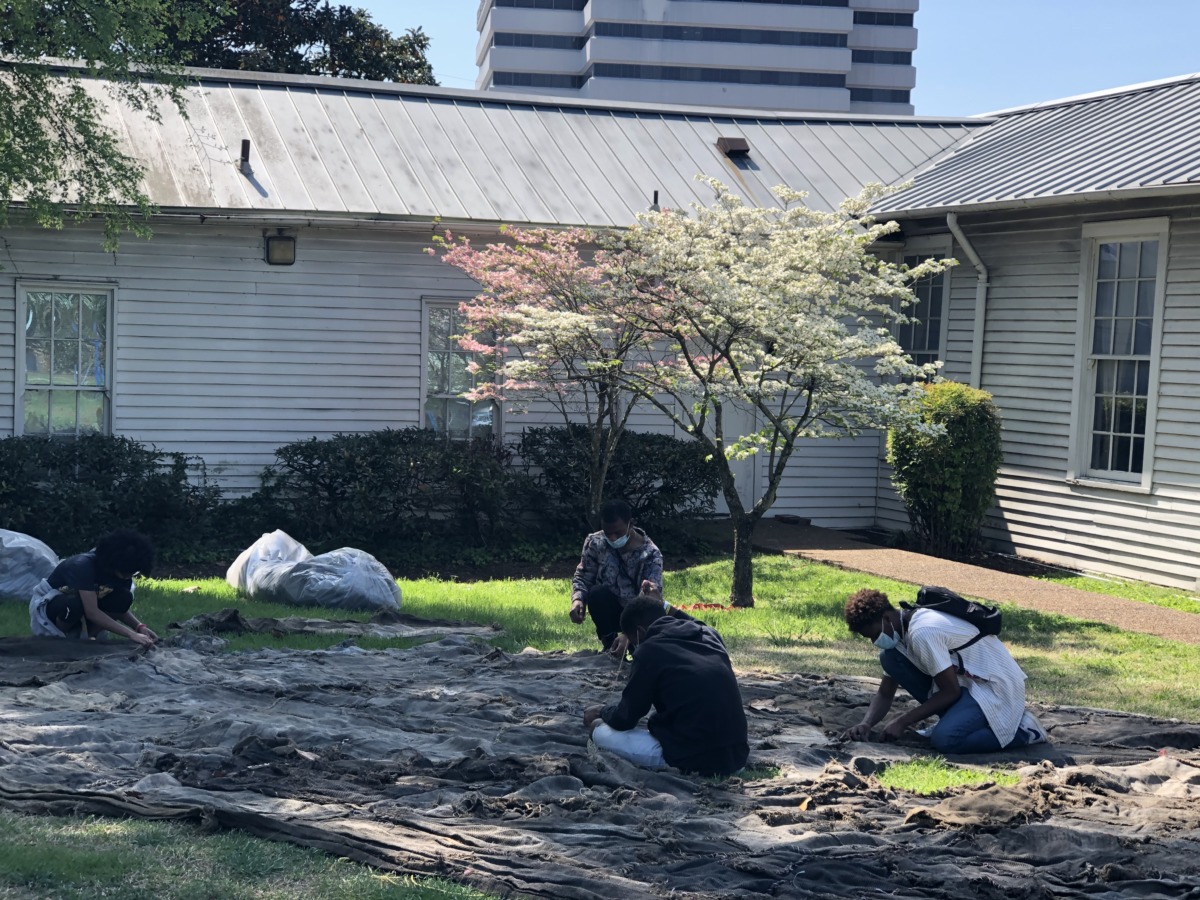 four people sit on the grass sewing burlap sacks. 