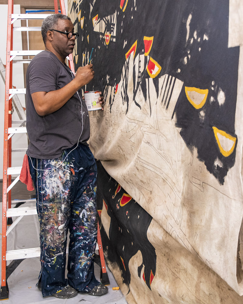 a Black man in a grey-black shirt and paint splattered jeans stands in front of a red and silver ladder while painting a very large beige canvas with black, red, and yellow, paint