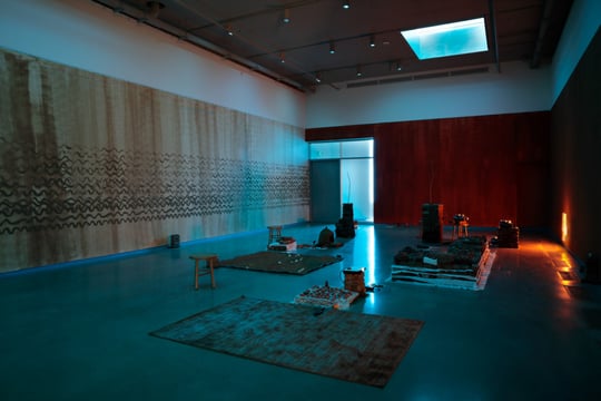 bright blue and rich red light wash over an exhibition space. brown lines swirl in horizontal waves on one wall, a small brown rug with similar patterns lay on the floor and the room is set like an interactive altar, stools and stones, set in piles surround blankets of dirt and white fabric
