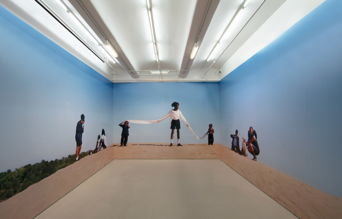 Installation view of a panoramic color photograph of three groups of Black people on a sand dune on a sunny day.