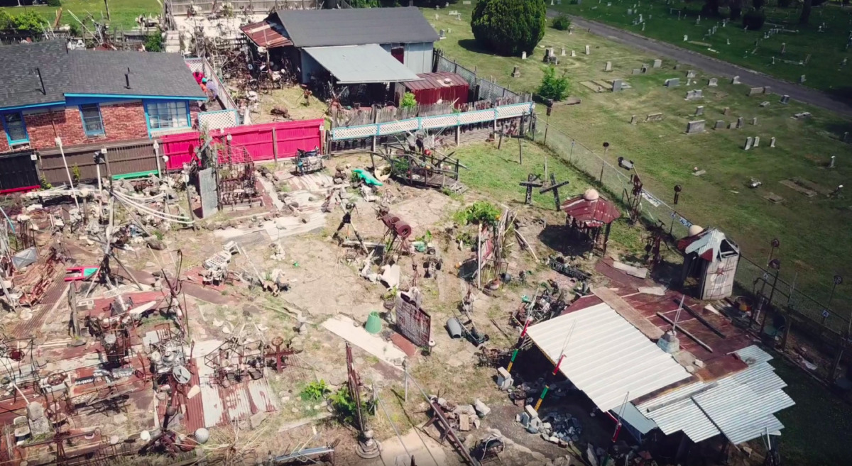 an aerial shot of a multi-acre yard show with collections of metal, painted signs, and sculptural artifacts in careful, yet overwhelming proportions, on the right side of the yard is a cemetery of green grass and humble gravestones