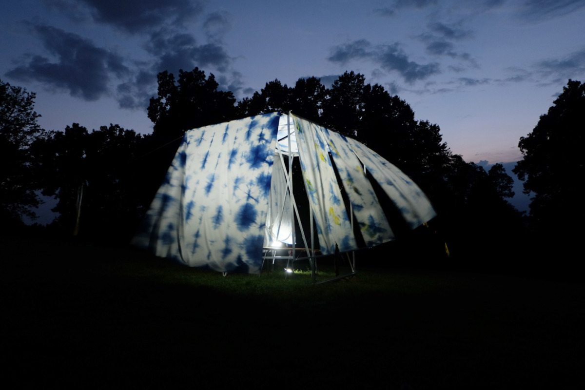 photograph of an outdoor tent made from white fabrics dyed with blue pigment, the wind has caught the fabric and the sheets are blowing in twilight, surrounded by a lavender cloud-kissed sky and the green-black silhouette of surrounding forest