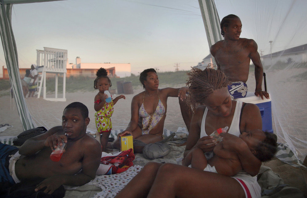 a Black family lounging on a beach at sunset, a few members are looking toward the direction of something, perhaps the setting sun. a small toddler drinks from a Hug fruit barrel and stares at the camera while sipping blue juice. in the foreground, a young mother holds her baby and feeds it a bottle