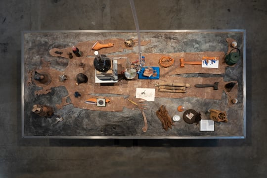 a tobacco leaf cut in the shape of a human body on a steel table. there are various objects on the silhouette including ceramics, clay, plants and herbs