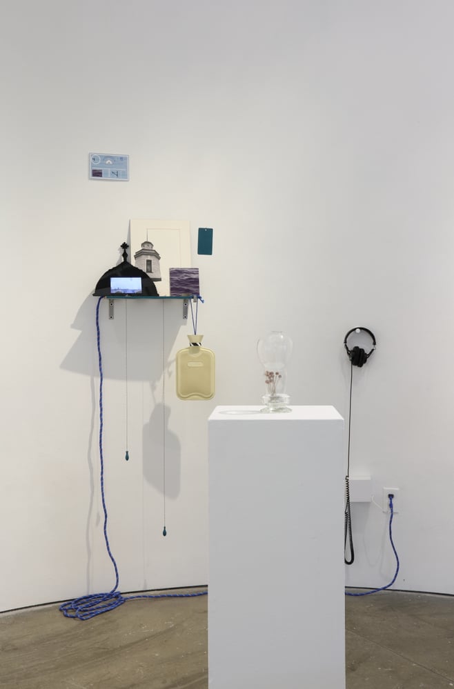 a white gallery with hanging objects including a pair of black headphones, hot water bottle, an extension chord, a picture of a church.