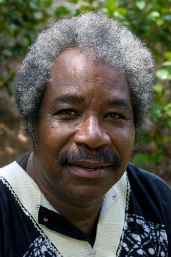 headshot of a Black man with a short salt and pepper afro, wears a white and black collared shirt against a blurred green outdoors background