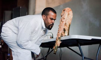 a Black man blows smoke at a horn that looks as though its been deep fried in grease, like a chicken wing. he wears a white jumpsuit