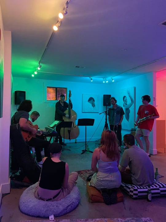 interior of a white gallery with multicolored ambient lights, a crowd surrounds a group of musicians playing jazz music in the dimly lit space