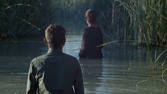 two boys waist deep in a watery marsh, turned with their clothed backs to the camera