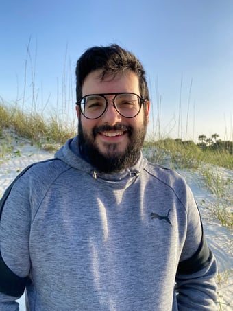 A headshot of Christopher, who is standing on a beach. He has thick black hair and a full beard that covers his light beige skin. He wears large circular glasses and a large smile on his face