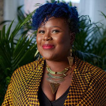 Courtney smiles in an interior scene, where she is backed by green plants. She wears red lipstick, and a fitted blazer with a checkered print. Her coily hair is dyed blue and sweeps her forehead