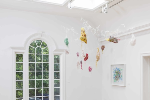 Mobile of brightly colored plastic sculptures with an ornately paneled window, where verdant evergreens can be seen outside the gallery