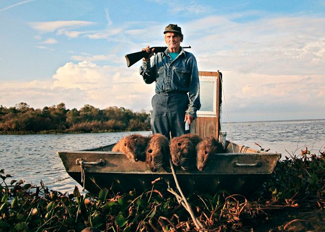 Man stands on a small boat with gun propped up on his shoulder annd four large rodents lay in front of him. 