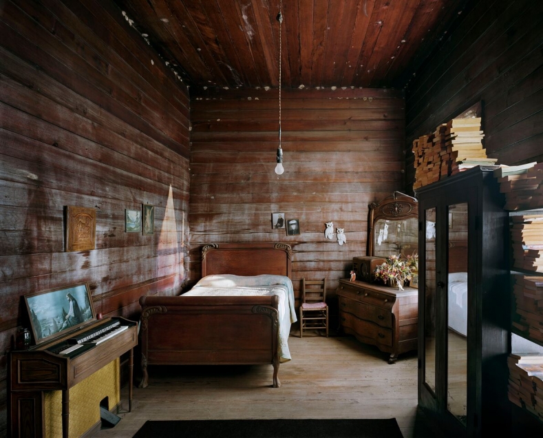 Image of a photograph by Andrew Moore showing the interior of a log cabin, a bed, books, and a small piano.