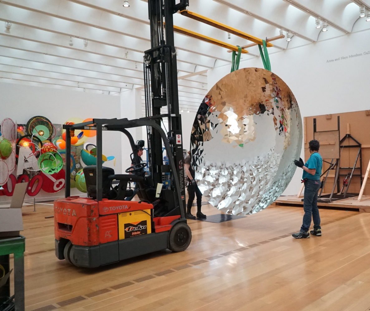 a crane moves kapoor's large round stainless steel sculpture that gives kaleidoscope-like reflection. a person stands next to the sculpture directing it and looks very small in comparison