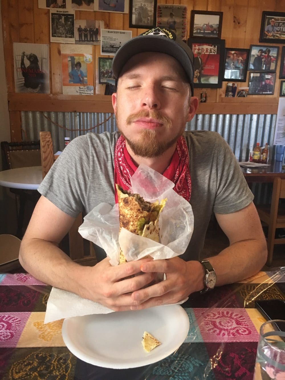 A fair complected man with facial hair, wearing a red bandana and gray t-shirt, holding a burrito in front of him with his eyes closed.