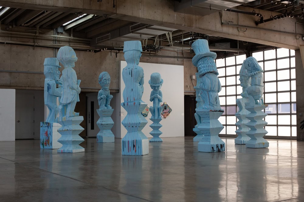 a large gallery with a concrete floor and wall of windows in the background, with seven greenish totem-like sculptures arranged in the center.
