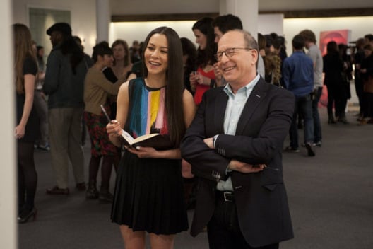 A woman holds an open book in her hands and a man stands next to her with crossed arms, both looking at something to the left.