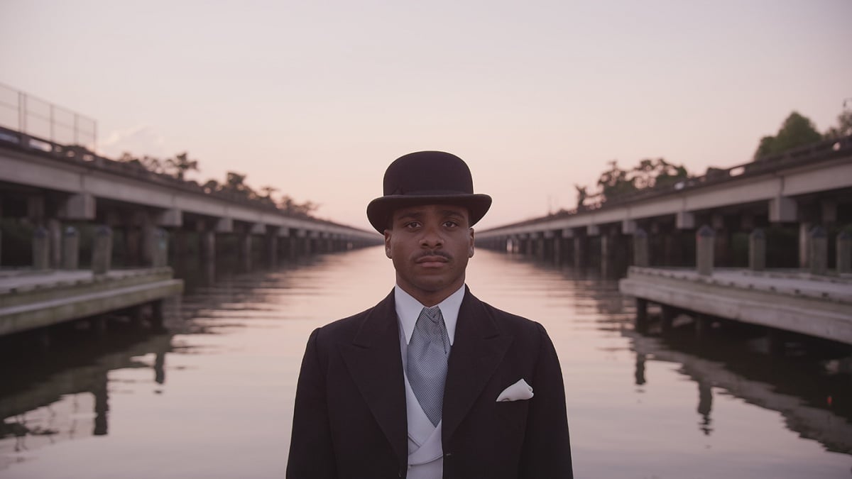 Black man in a bowler hat and suit, with a water canal receding in the background, from a John Akomfrah film called Precarity. 