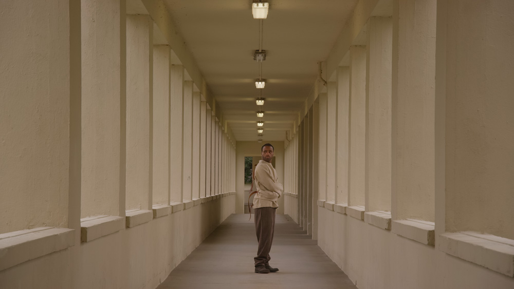 Black man in a strait jacket standing in a corridor looking at viewer. From John Akomfrah film Precarity. 