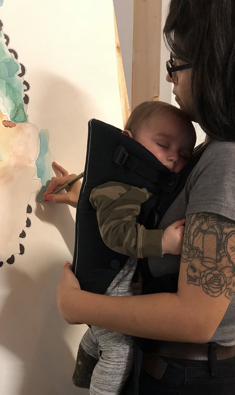 A woman with tattoo, holding her sleeping baby in a baby carrier as she works on a painting.