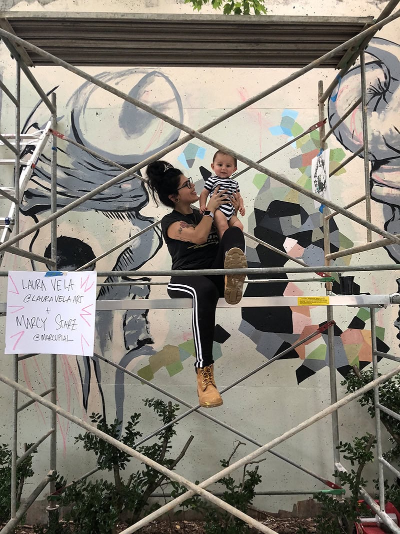 Woman sitting on scaffolding in front of a mural and holding her baby up.