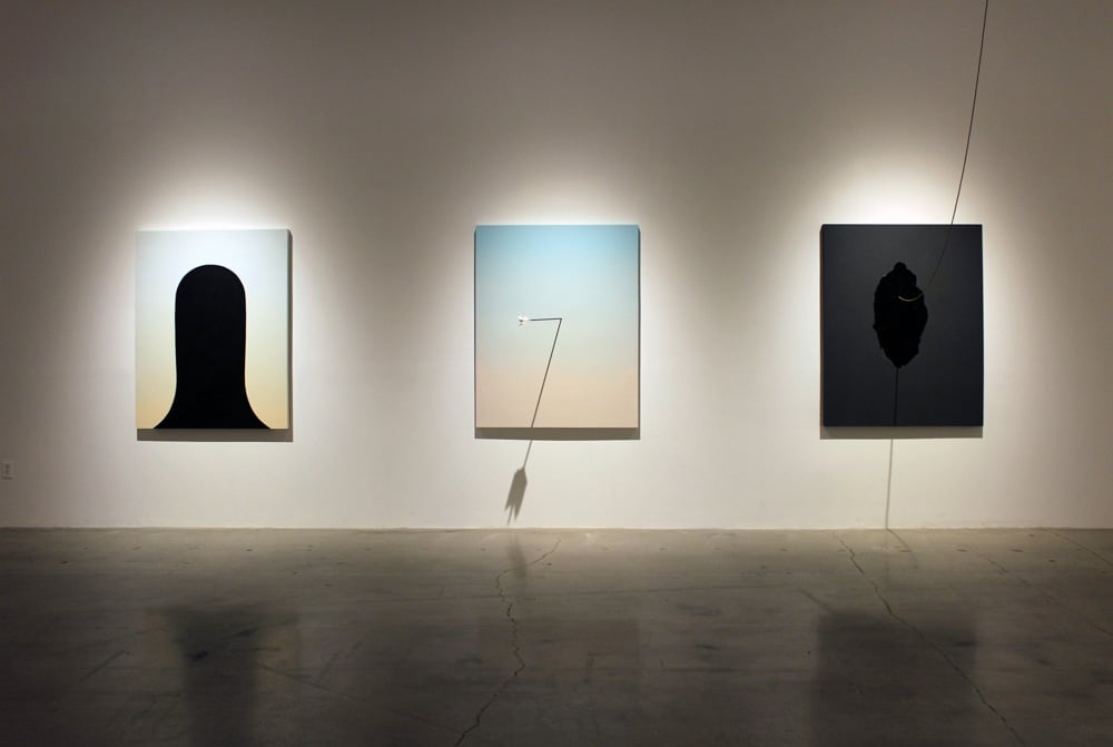 Three paintings by Kirstin MItchell. From left, light background with headlike silhouette, pastel colorfield with an actual arrow piercing the surface, and a dark background with a dark center and an actual cord that stretches up to the ceiling.