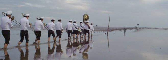 A line of 10 musicians marching in a flooded street with a tuba leading the way, seen from behind. 