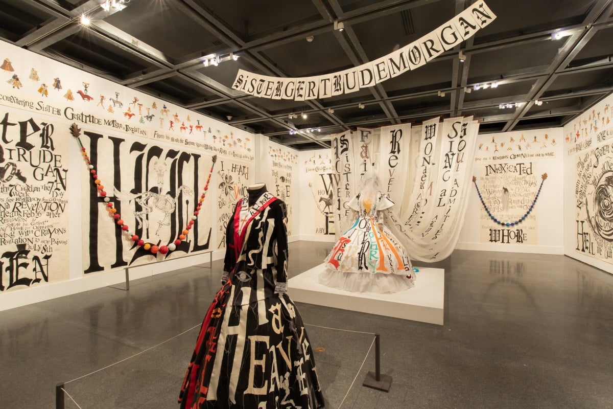 A gallery-filling installation with a headless mannequin in the foreground wearing a long black dress, another in the background in a flowing white dress, and text and images all over the walls, by artist Lewley Dill at the New Orleans Museum of Art.