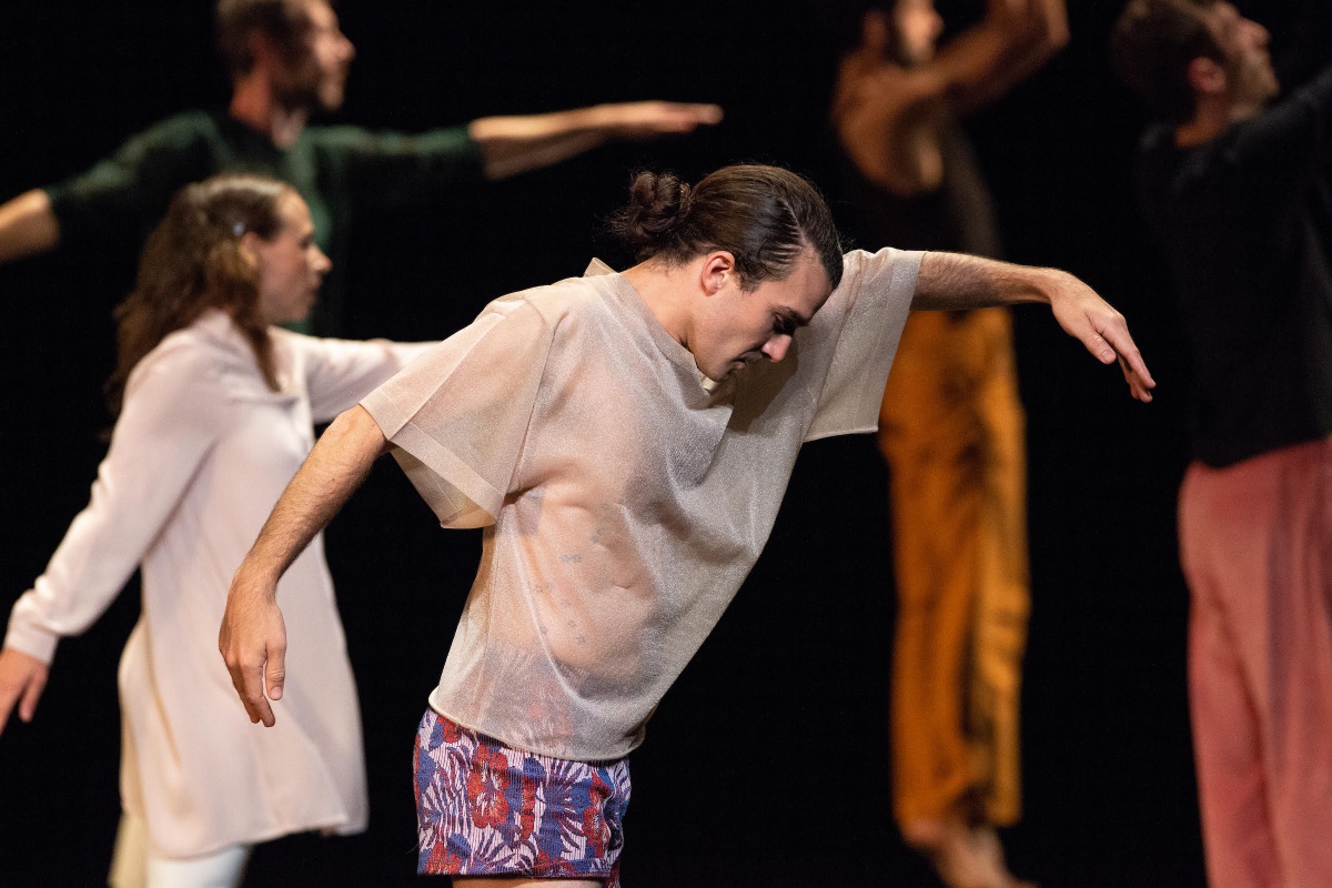 A man facing sideways with arms lifted, and more dancers in the background, a scene from Long Run performance choreographed by Tere O'Connor.