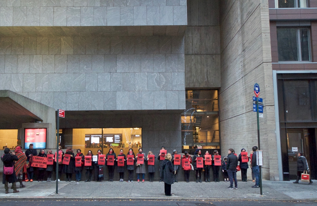 A long row of #metoo protestors holding red signs in front of the Met Breuer building. 