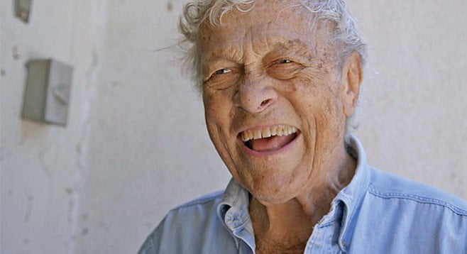 Head shot of an elderly man, laughing, in a blue shirt and messy gray hair.