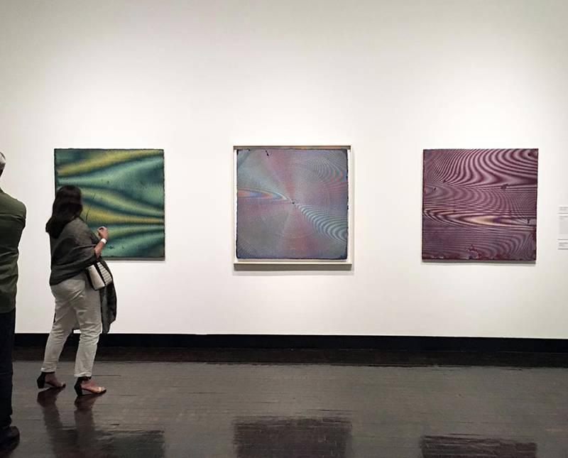 Three abstract monochromatic paintings on a wall with a woman standing to the left looking at them. included in Chaos and Awe at Frist Art Museum