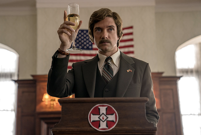 A white man holding up a champagne glass standing at a podium with KKK symbol on front and and American flag on the wall behind him