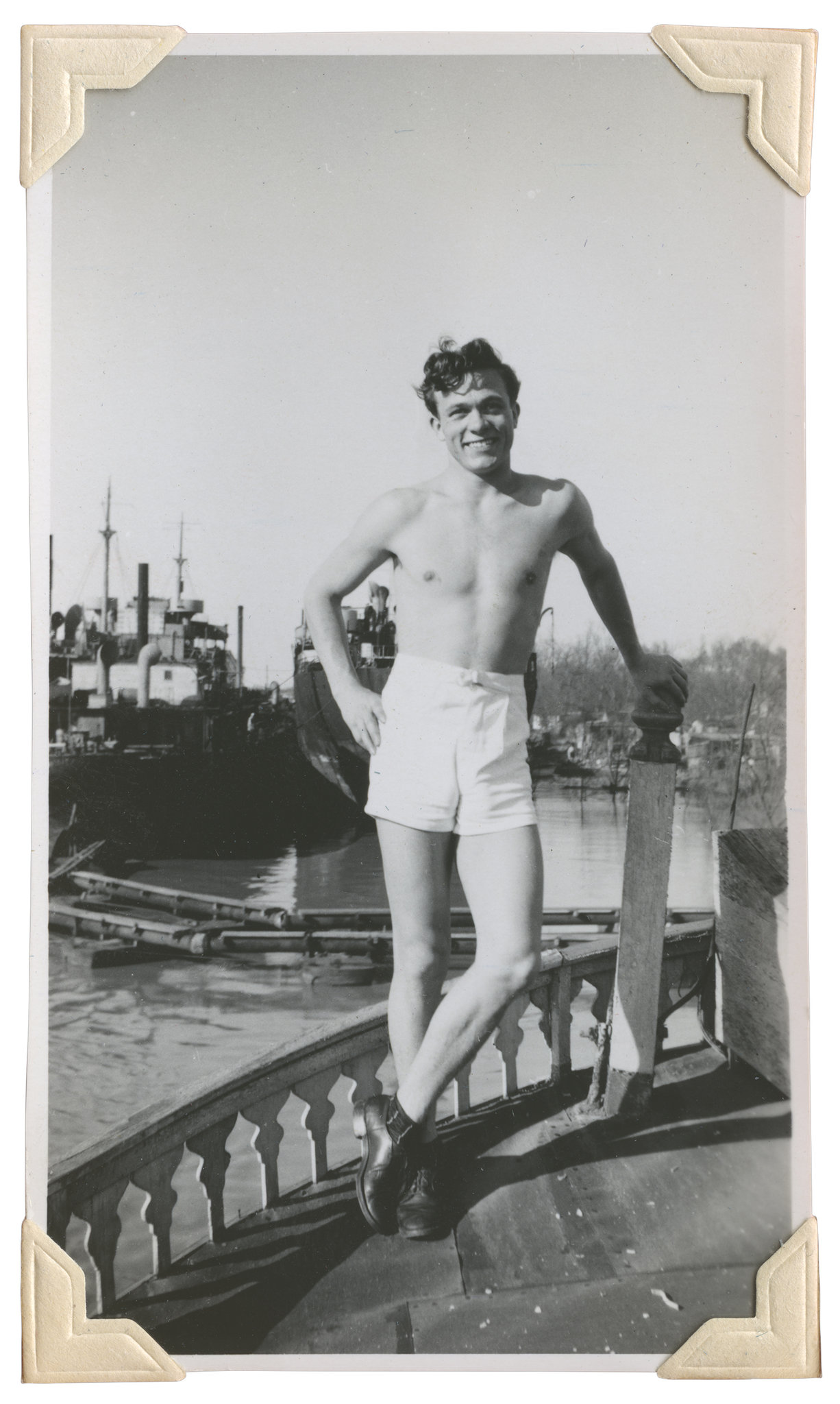 Vintage black and white photo of a slender shirtless man in shorts leaning on a fence post.