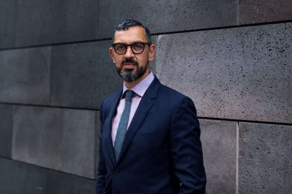 An adult man with a beard wears glasses and a suit and tie in front of a gray stone wall.