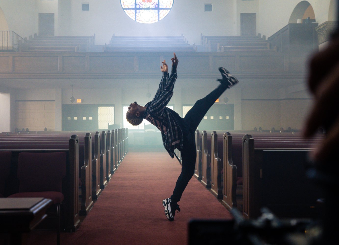 a young black man in a plaid shirt, dark pants and tennis shoes dances in-between church pews.
