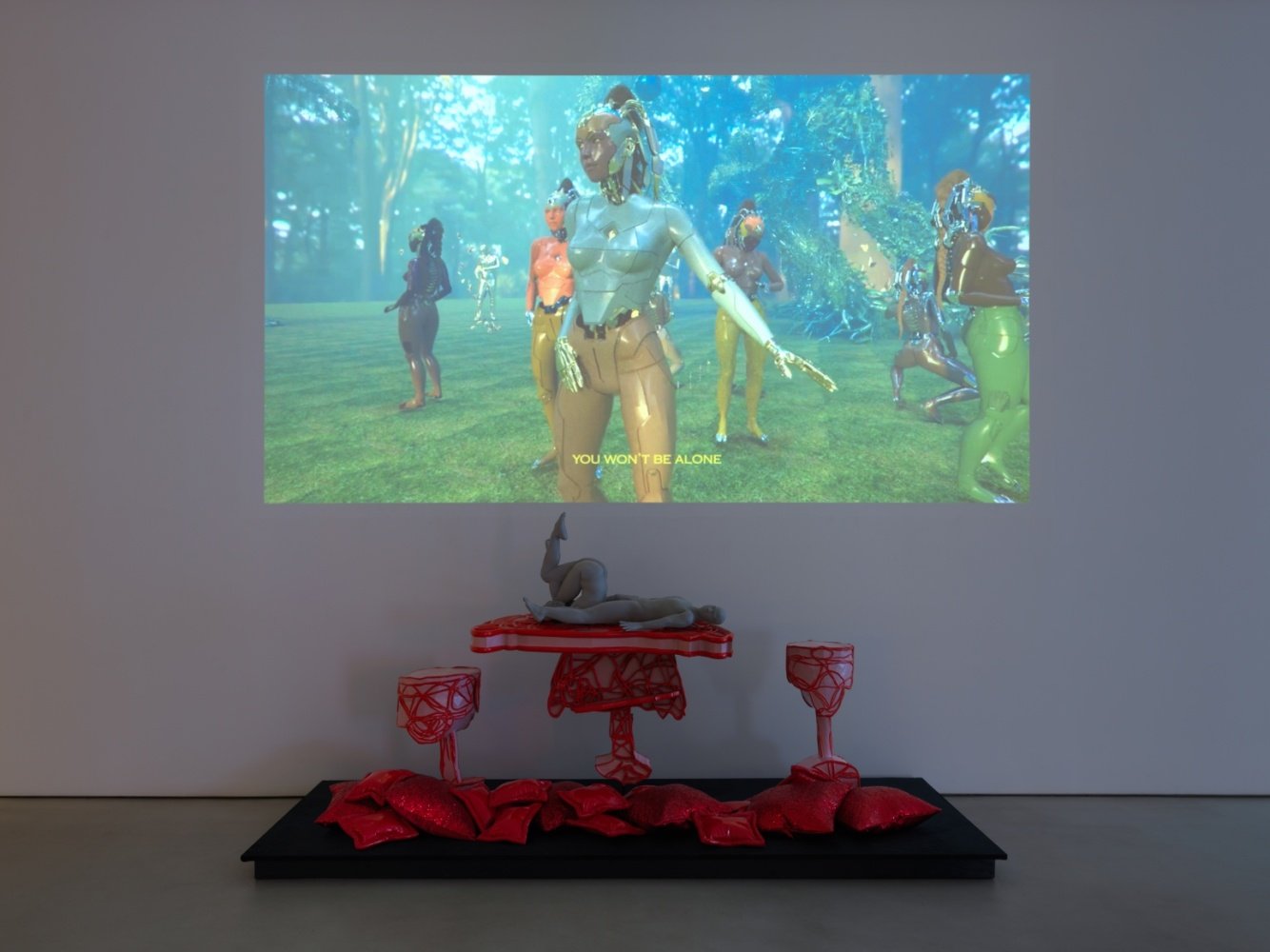 a video still of black robots above a sculpture of figures reclining on red pillows