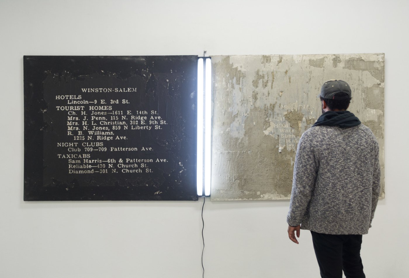 a man stands in front of two painted canvas with a fluorescent bulb between them, one white and one black with text about black friendly lodging. 