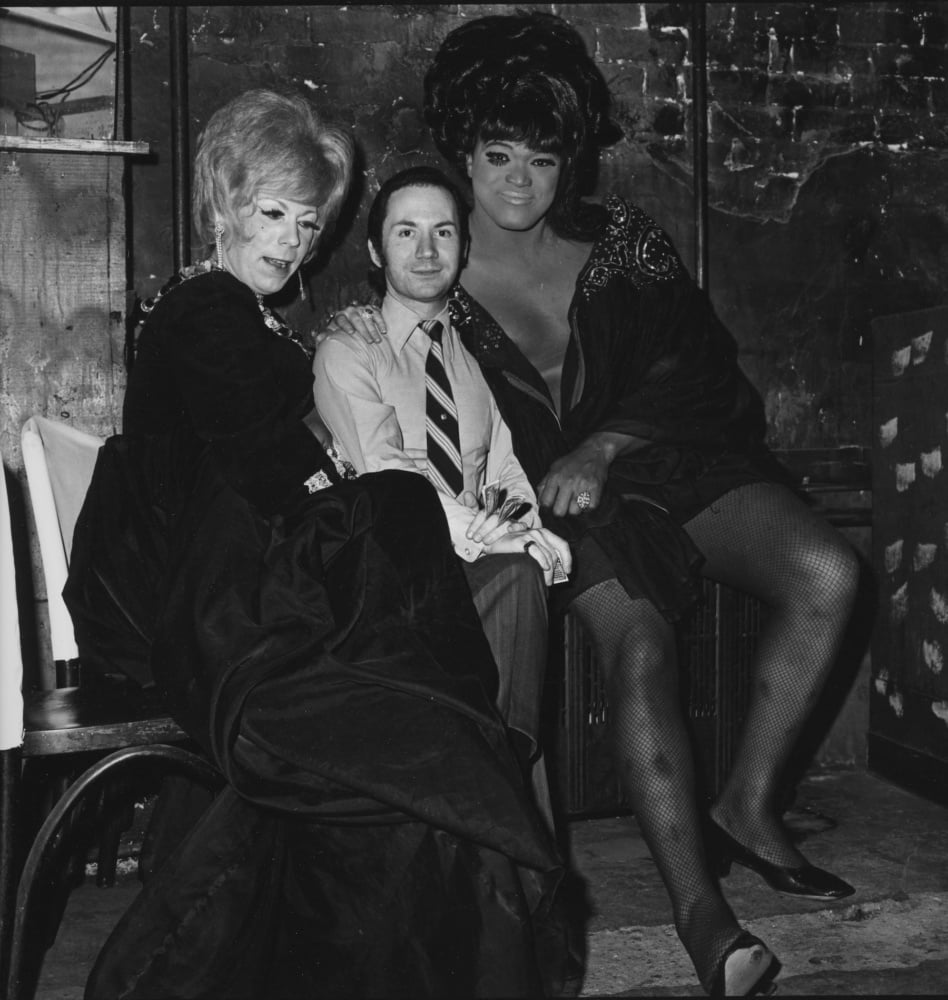 a black and white image of two drag queens embracing a man in a tie.