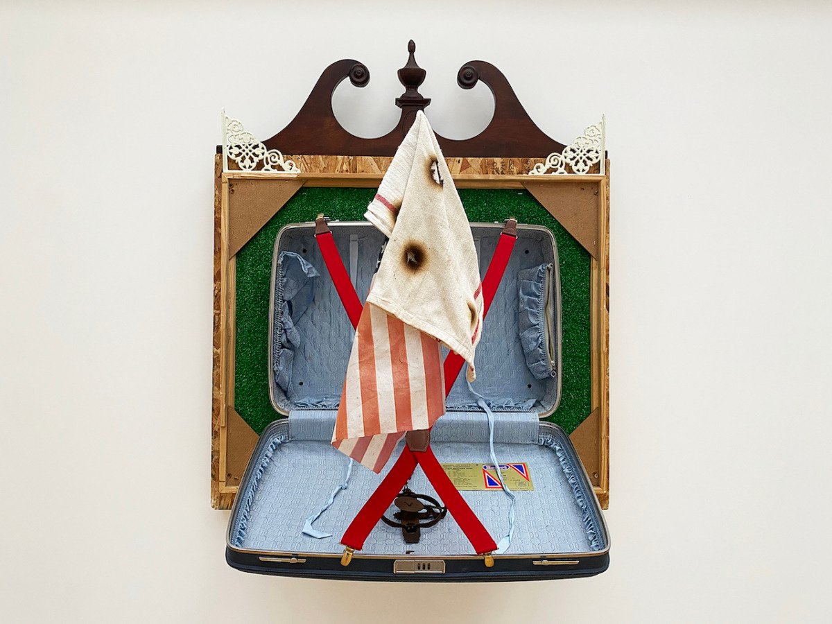 a suitcase mounted on the wall is hanging open with red suspenders, a burned towel and an American flag inside it. 