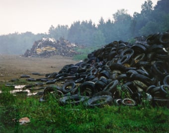 A large pile of discarded black tires lie in the foreground of a trash heap in a dumping ground.