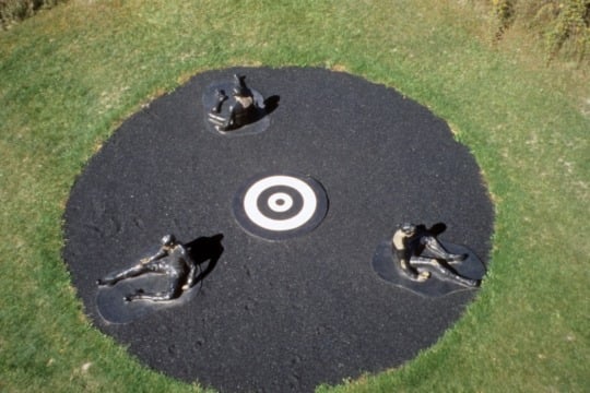 an aerial view of an outdoor installation, a large charcoal circle encompasses a concentric white bulls-eye, three life-size sculptures of a Black man in a black jumpsuit sit equidistant from each other along the circle's edge. the circle is surrunded by a patchy grass field