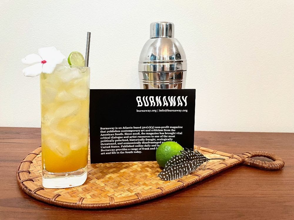 a golden cocktail in a highball glass with a white flower next to a cocktail shaker, green lime and black postcard with text