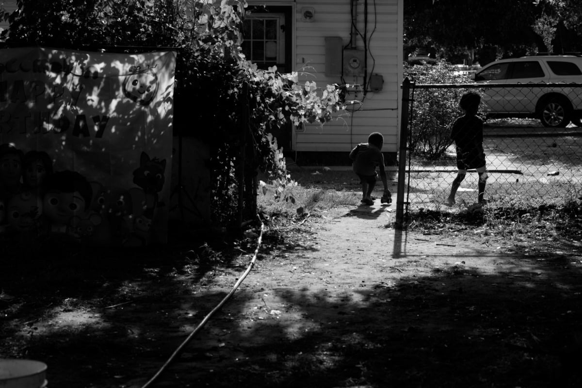 black and white photograph of two small children behind a house with an open wire gate. the contrast is high and not many details can be made out, but the shadows cast a playful and striking scene with cascading leave patterns from the tall trees above