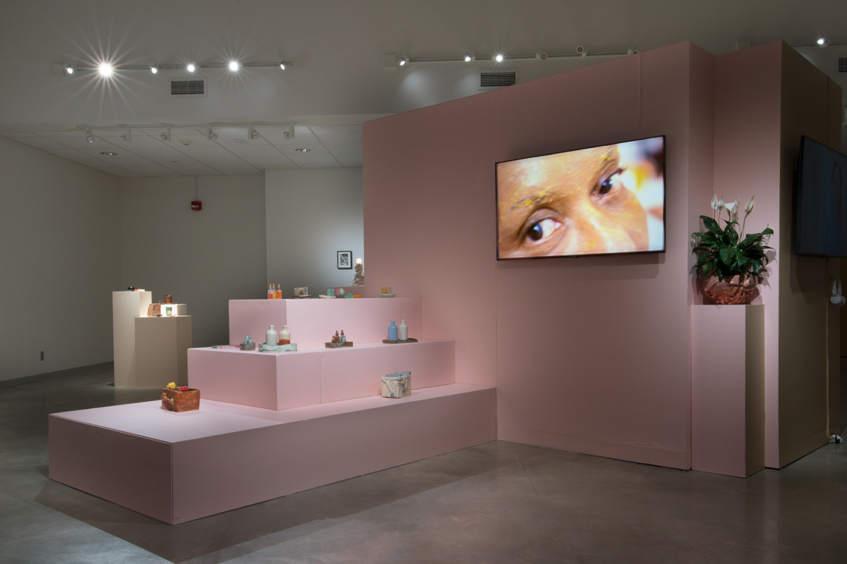 installation view of sculptures on pink blocks, a video screen with a still of a black woman's eyes