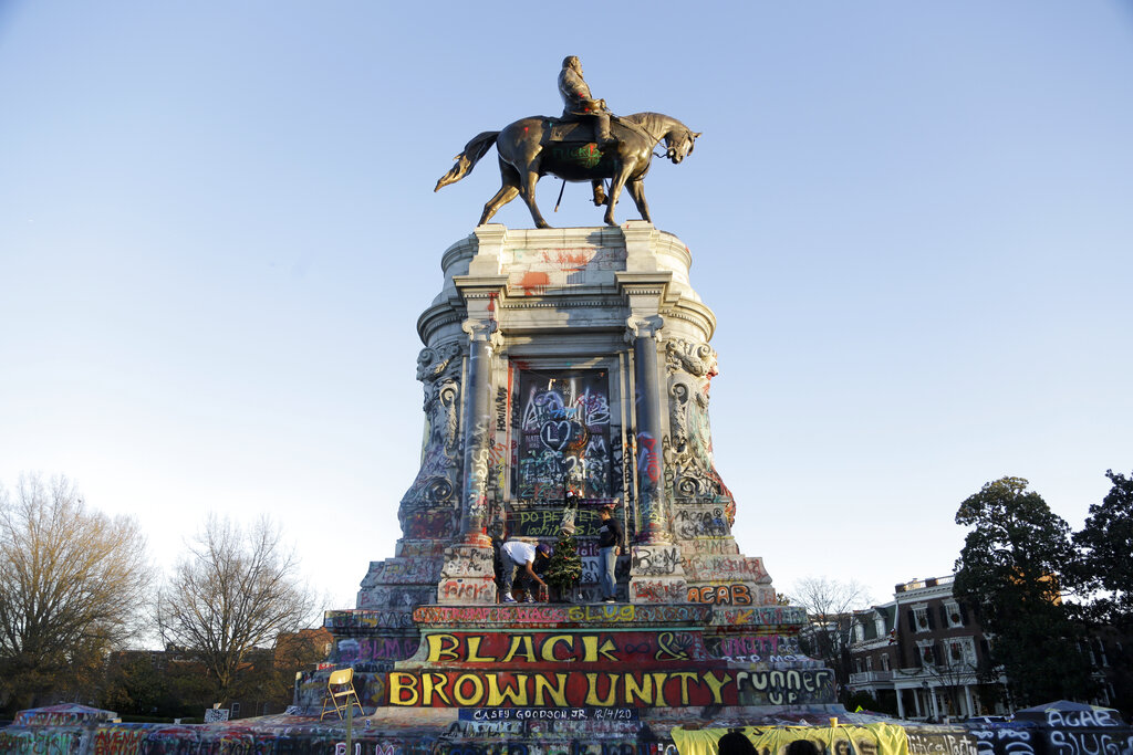 Two people of color decorate a Christmas tree placed on the base of the Gen. Robert E. Lee statue, covered in vibrant graffiti with phrases like "BLACK AND BROWN UNITY" and "TRUMP IS WACK."