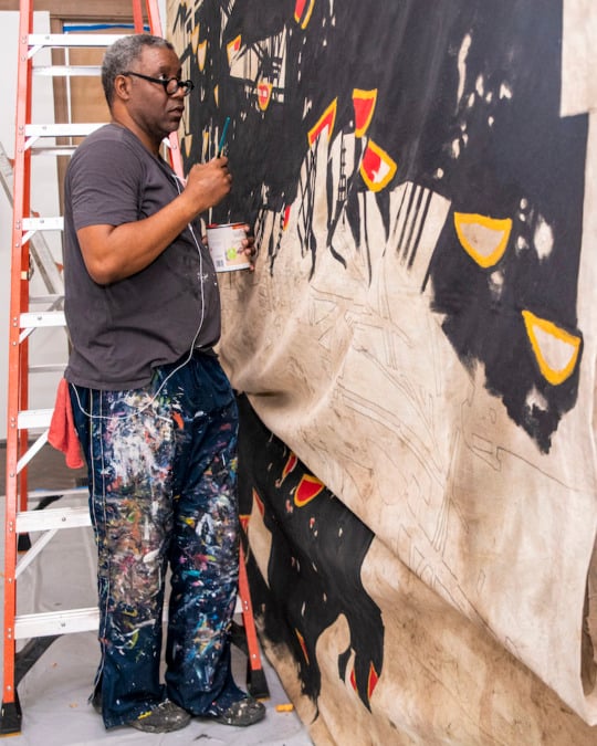 a Black man in a grey-black shirt and paint splattered jeans stands in front of a red and silver ladder while painting a very large beige canvas with black, red, and yellow, paint